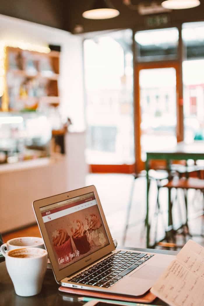 A laptop displaying a website sits on a table with two coffee cups in a cozy cafe environment.