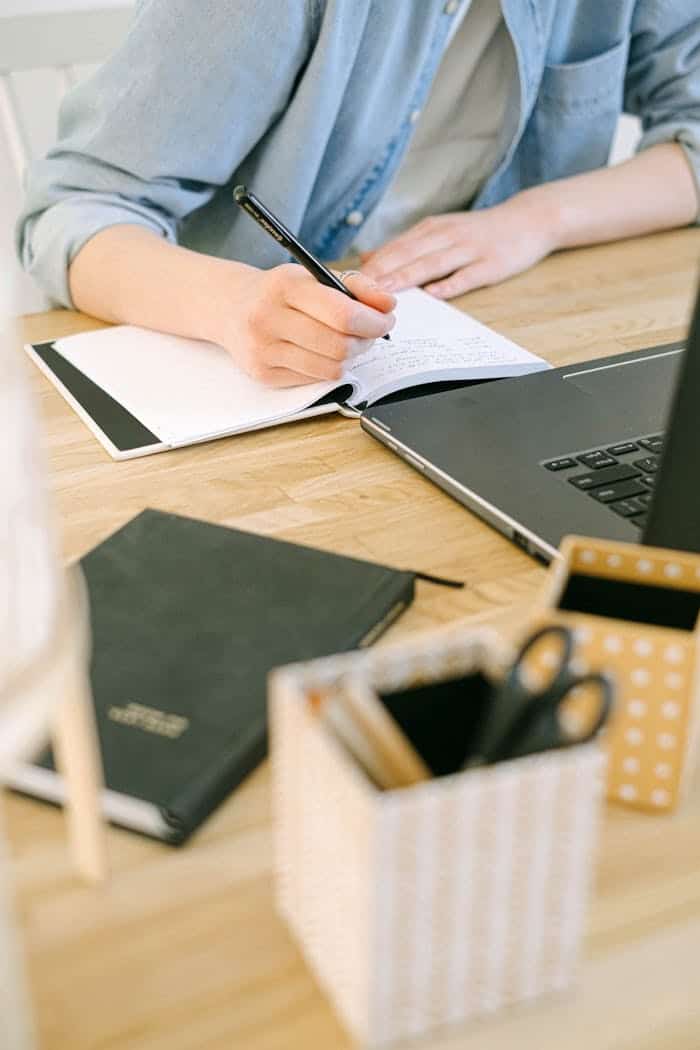 Person writing in notebook with laptop and stationery on desk.